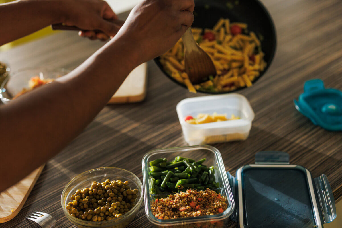 Your Gut Already Makes Its Own Ozempic — Here's How to Boost It! - 3 A person serves pasta from a pan into a container on a table with bowls of green beans, peas, and another glass container with food—creating a balanced meal ideal for supporting gut health.
