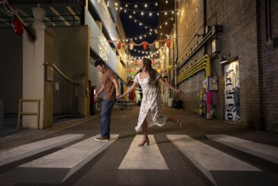 A woman in a white dress playfully balances on a crosswalk while a man in jeans and a brown shirt walks ahead in a lit urban alley at night—perfect inspiration if you visit Hattiesburg, MS for unique things to do.