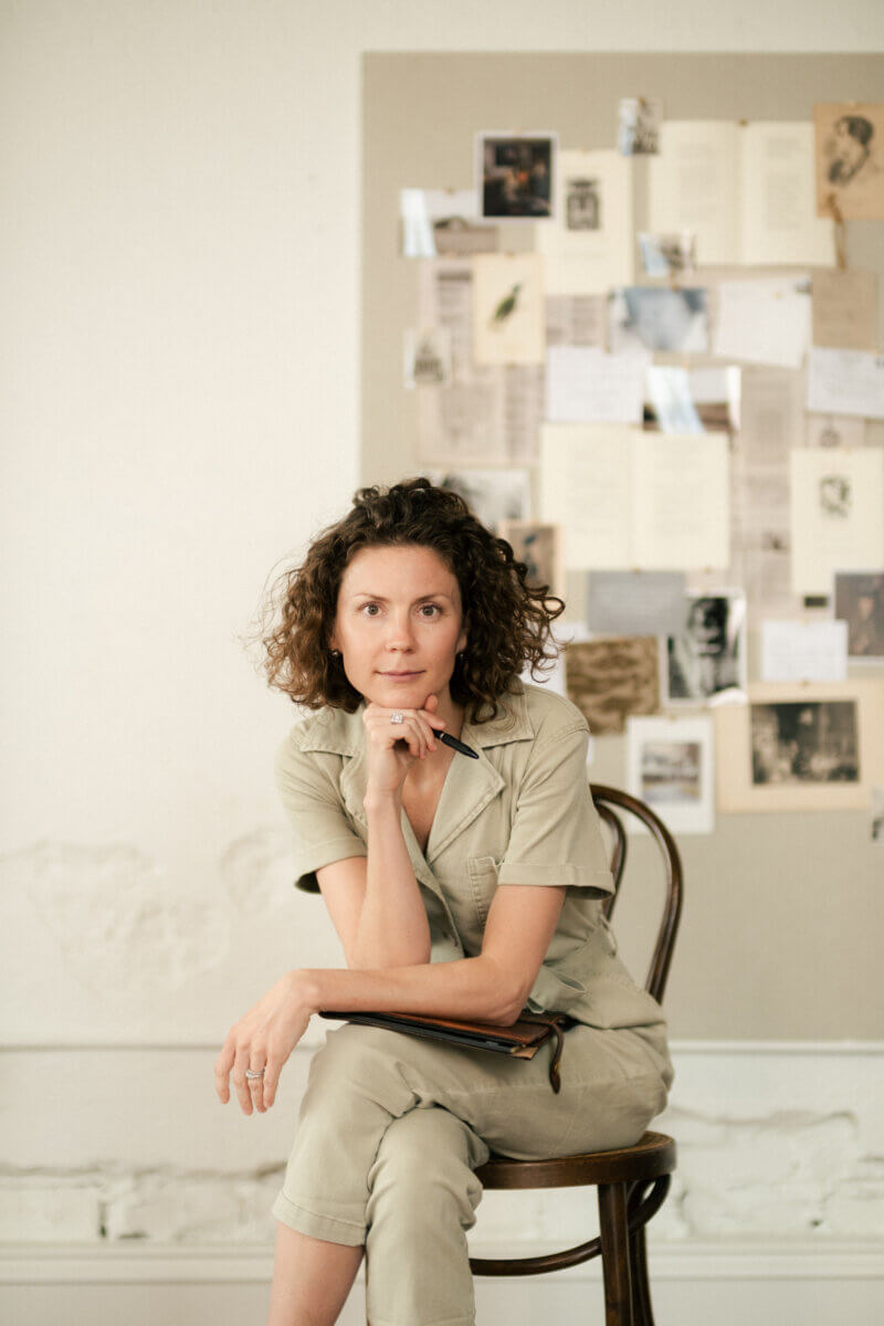 Inside Interior Designer Jen Bienvenu’s Wildly Collected World - 2 A woman with curly hair sits on a chair, holding a notebook and pen, in front of a bulletin board covered with papers and photographs.