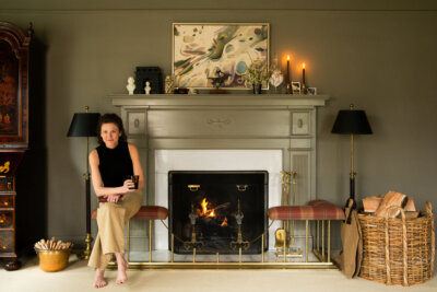 A woman sits on a bench by a lit fireplace in a living room, surrounded by candles, artwork, and baskets of firewood—a cozy scene where design inspired by humanity meets the natural world.