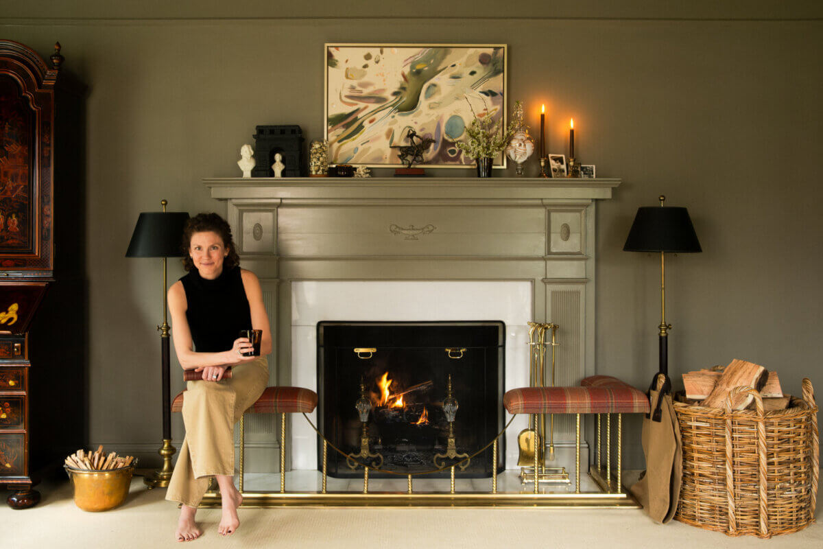 A woman sits on a bench by a lit fireplace in a living room, surrounded by candles, artwork, and baskets of firewood—a cozy scene where design inspired by humanity meets the natural world.