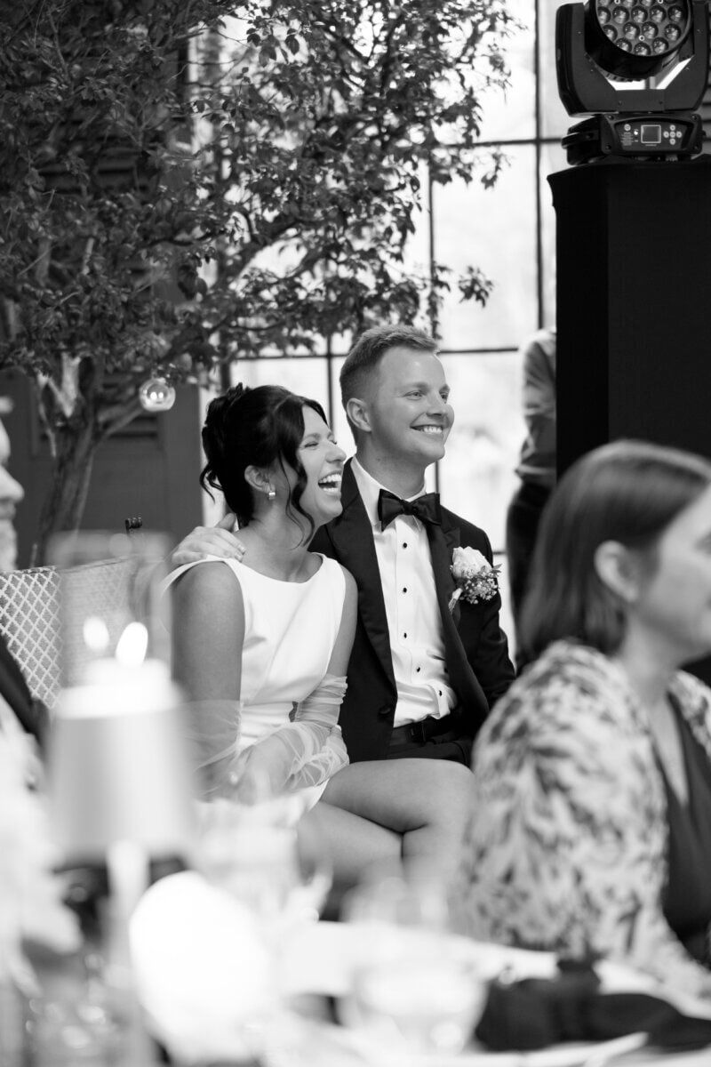 A bride and groom sit closely together, smiling as they watch something off-camera at their wedding reception.