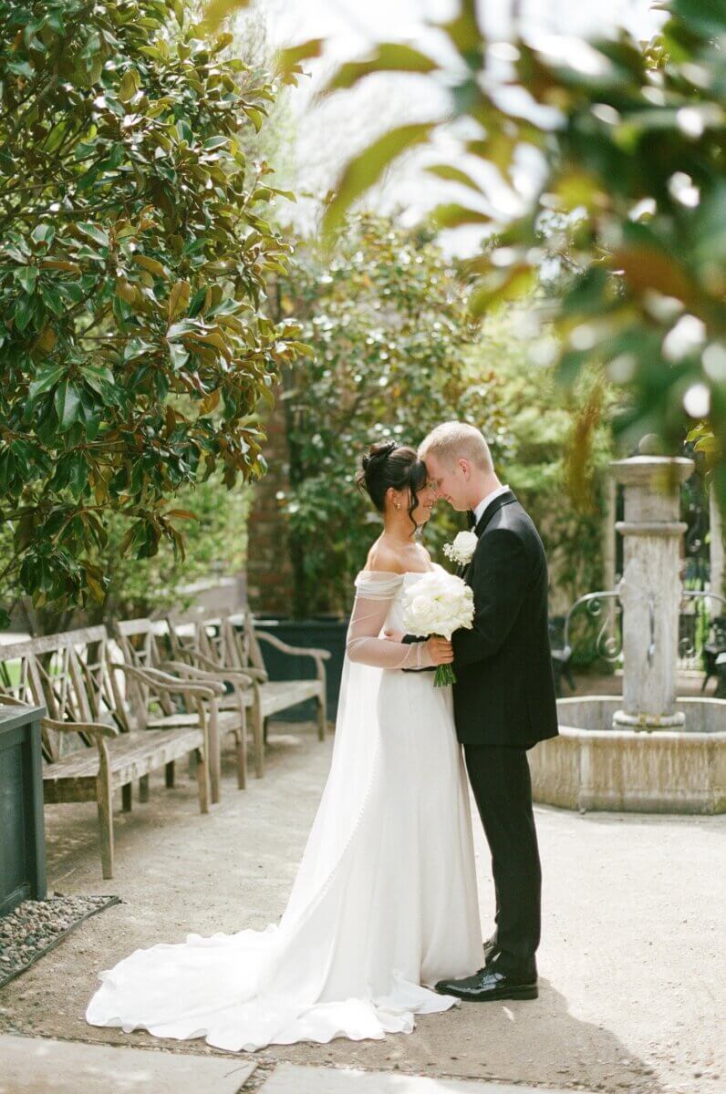 Disco Balls and Black-Tie Style Defined This Lexington Wedding - 2 A bride and groom stand close together outdoors, holding hands and a bouquet of white flowers, surrounded by greenery and benches near a stone fountain as they prepare to exchange heartfelt wedding vows.