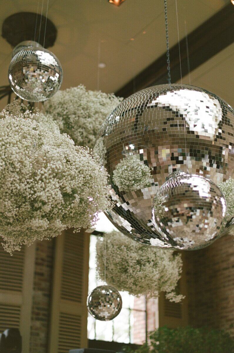 Several mirrored disco balls of varying sizes are suspended among large clusters of white baby's breath flowers indoors, creating a whimsical scene perfect for exchanging wedding vows.
