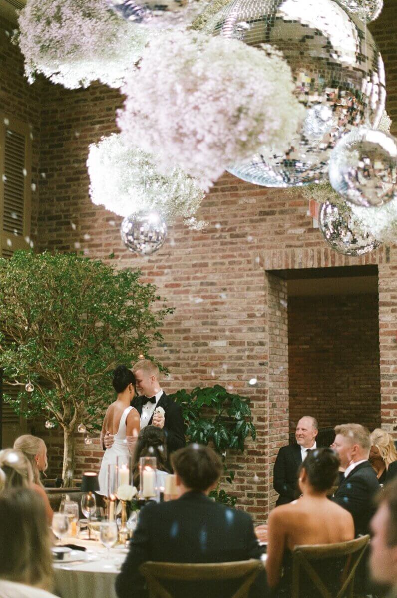 Disco Balls and Black-Tie Style Defined This Lexington Wedding - 6 A couple shares a dance at a wedding reception, surrounded by seated guests, as disco balls and floral arrangements hang from the ceiling.