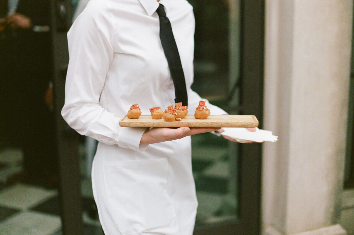 Disco Balls and Black-Tie Style Defined This Lexington Wedding - 9 A server in a white shirt and black tie holds a wooden tray with bite-sized appetizers in an elegant apiary, creating a formal setting perfect for exchanging wedding vows.