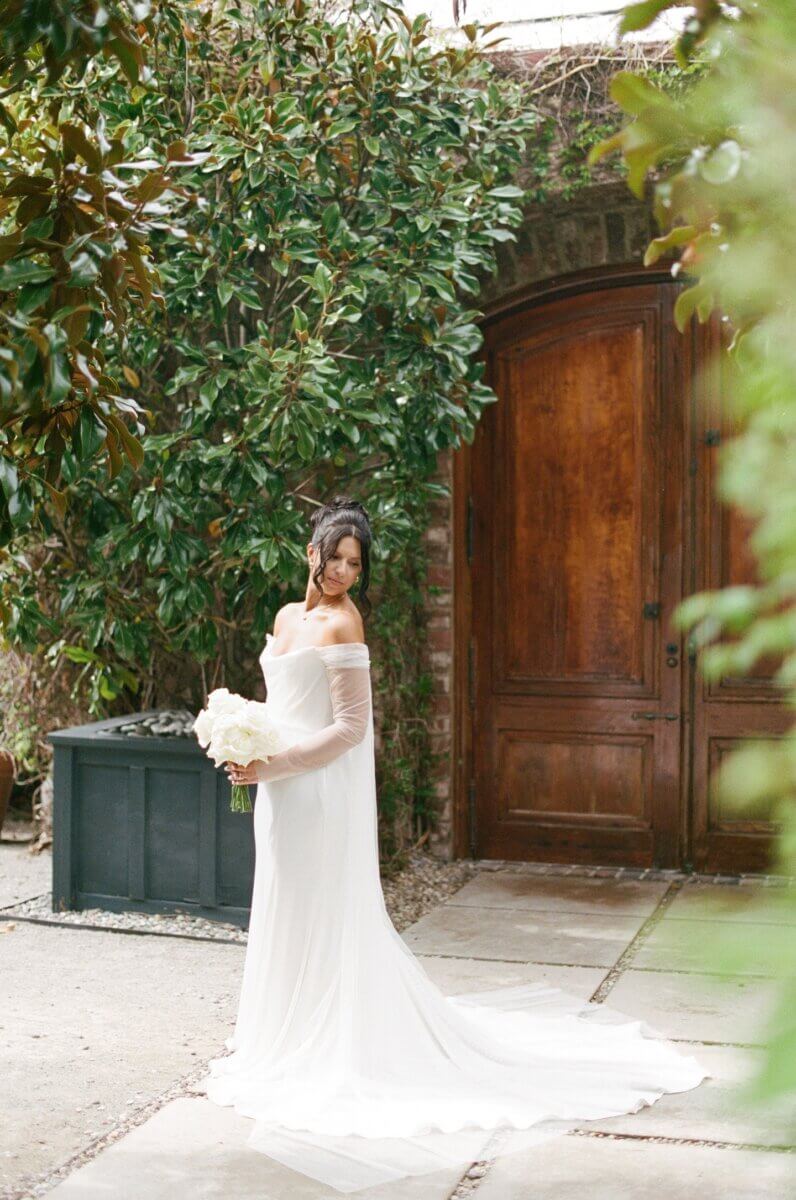 Disco Balls and Black-Tie Style Defined This Lexington Wedding - 4 A woman in an off-shoulder white wedding dress holding a bouquet stands near a wooden door and greenery, ready to exchange wedding vows in a charming outdoor setting.