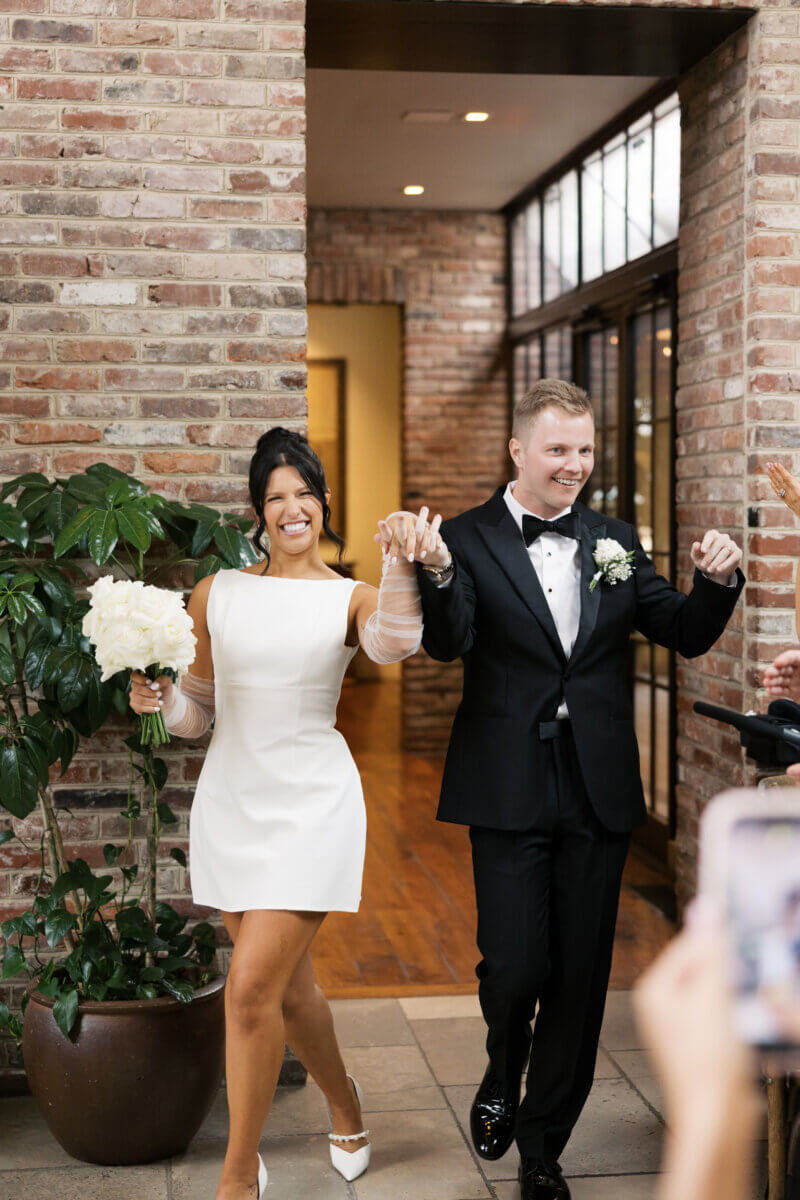 Disco Balls and Black-Tie Style Defined This Lexington Wedding - 10 A smiling couple in formal attire walks hand in hand indoors at their elegant wedding; the woman wears a white dress and holds a bouquet, while the man wears a black tuxedo.