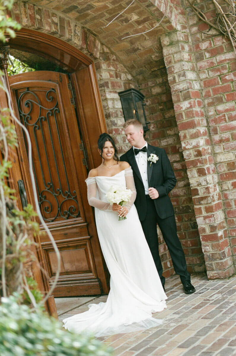 Disco Balls and Black-Tie Style Defined This Lexington Wedding - 3 A bride in a white gown and a groom in a black tuxedo stand together outside a large wooden door set in a brick wall, holding a white bouquet, ready to exchange their wedding vows.
