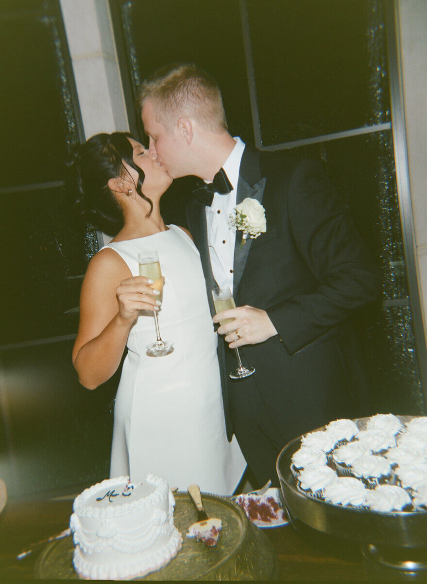 A bride and groom in formal attire share a kiss after exchanging wedding vows, holding champagne glasses beside a small wedding cake and a tray of cupcakes.