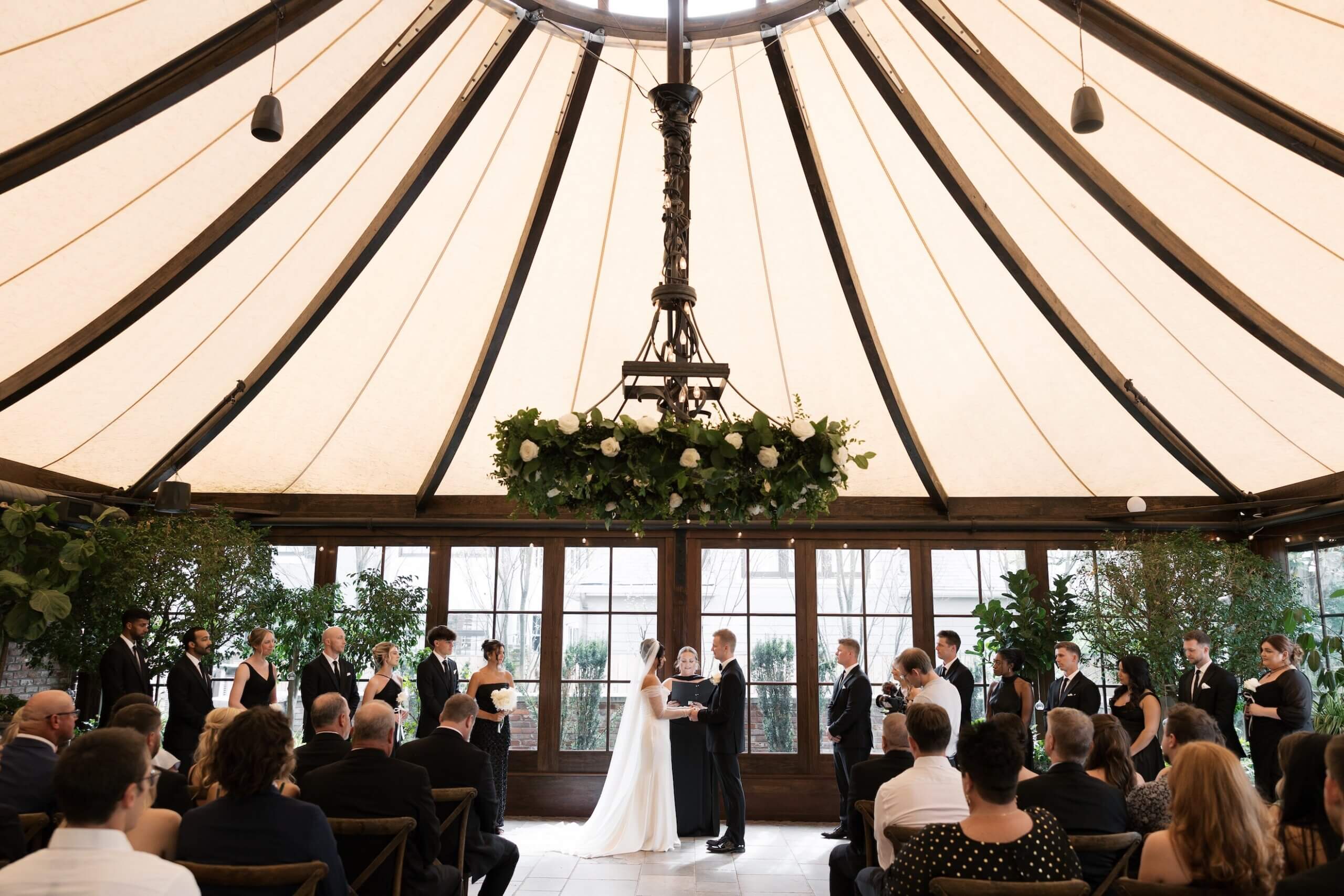A bride and groom stand at the altar under a large chandelier during an indoor wedding ceremony, surrounded by their wedding party and guests.