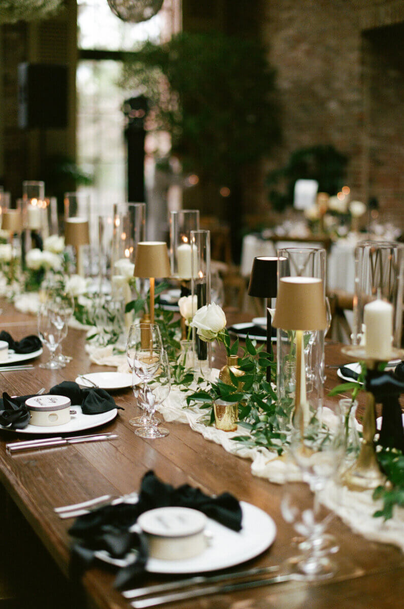 A wooden table is set for a formal event with white plates, black napkins, glassware, candles, and greenery as centerpiece decorations.