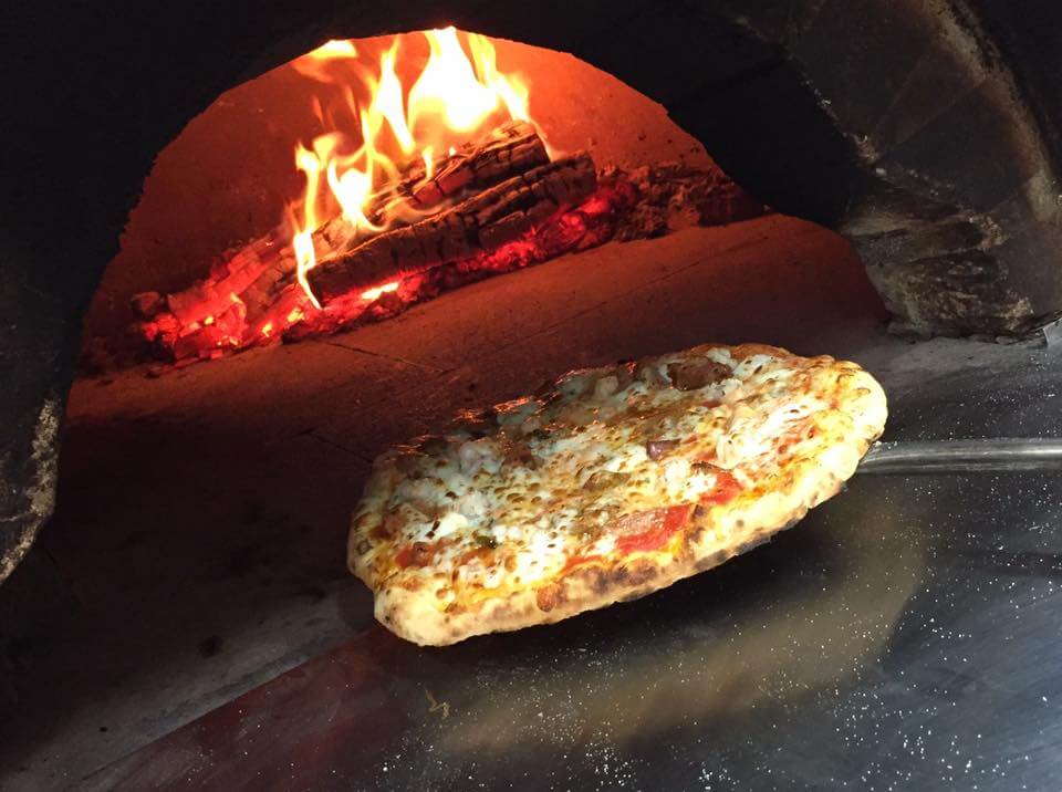 Savoring Louisiana: A Culinary Roadshow Across the State - 5 A pizza on a metal peel is being placed into a wood-fired oven with visible flames and burning logs, bringing true Louisiana culinary flair to life.