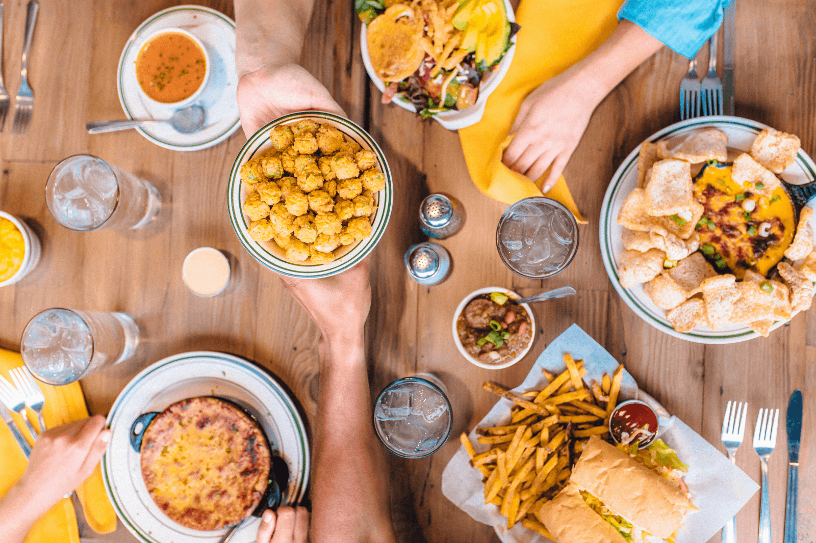 Overhead view of a table with various culinary dishes including fries, soup, salad, sandwich, casserole, and a bowl of fried bites—perfect for a Louisiana roadshow—with hands reaching for food and drinks.