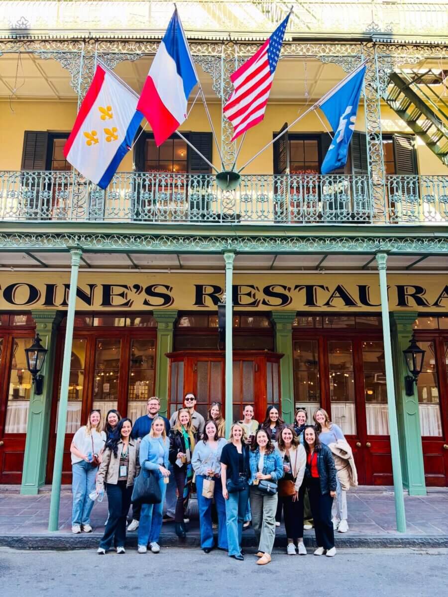 Savoring Louisiana: A Culinary Roadshow Across the State - 11 A group of people pose for a photo in front of a Louisiana restaurant, known for its rich culinary heritage, with four flags displayed above the entrance.