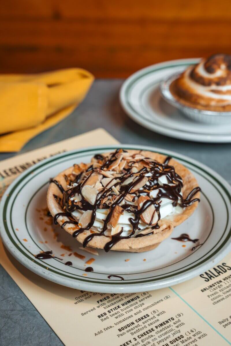 Savoring Louisiana: A Culinary Roadshow Across the State - 2 A small pie topped with whipped cream, chocolate drizzle, and sliced almonds sits on a plate next to a menu—offering a taste of the sweet treats often found in Louisiana restaurants—with a cinnamon roll in the background.
