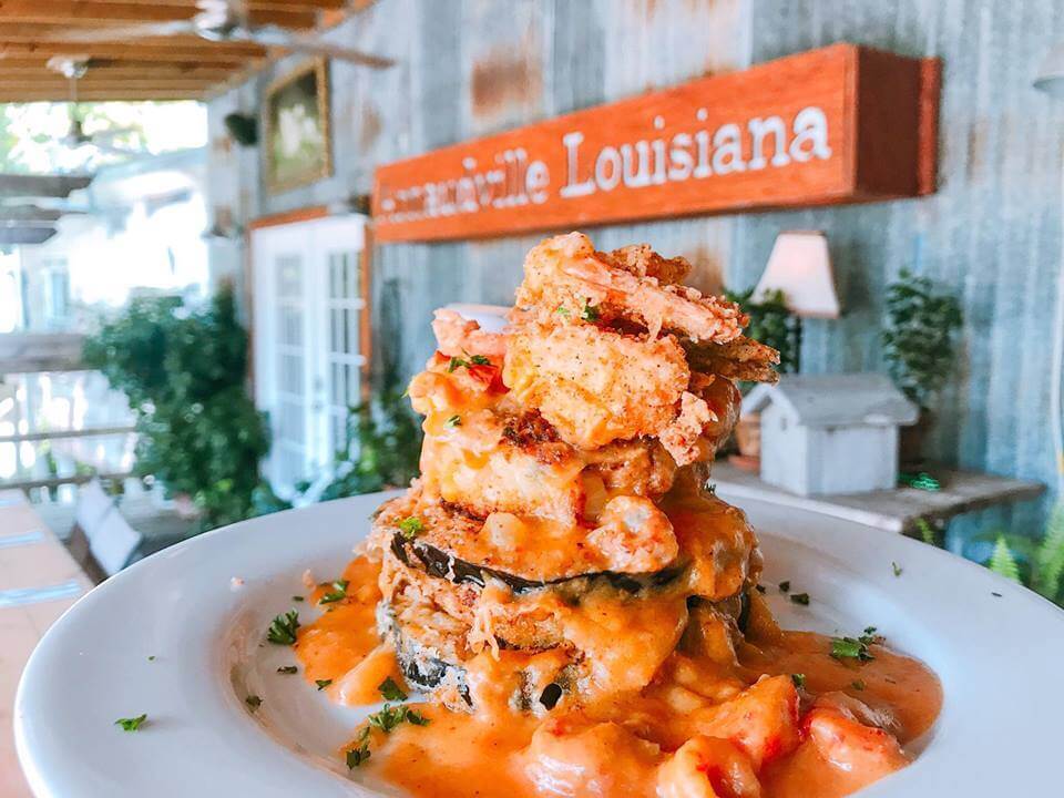 Savoring Louisiana: A Culinary Roadshow Across the State - 4 A plate with a stack of fried seafood, vegetables, and creamy sauce is served in a rustic restaurant with a “Louisiana” sign in the background, capturing the flavors of Louisiana.