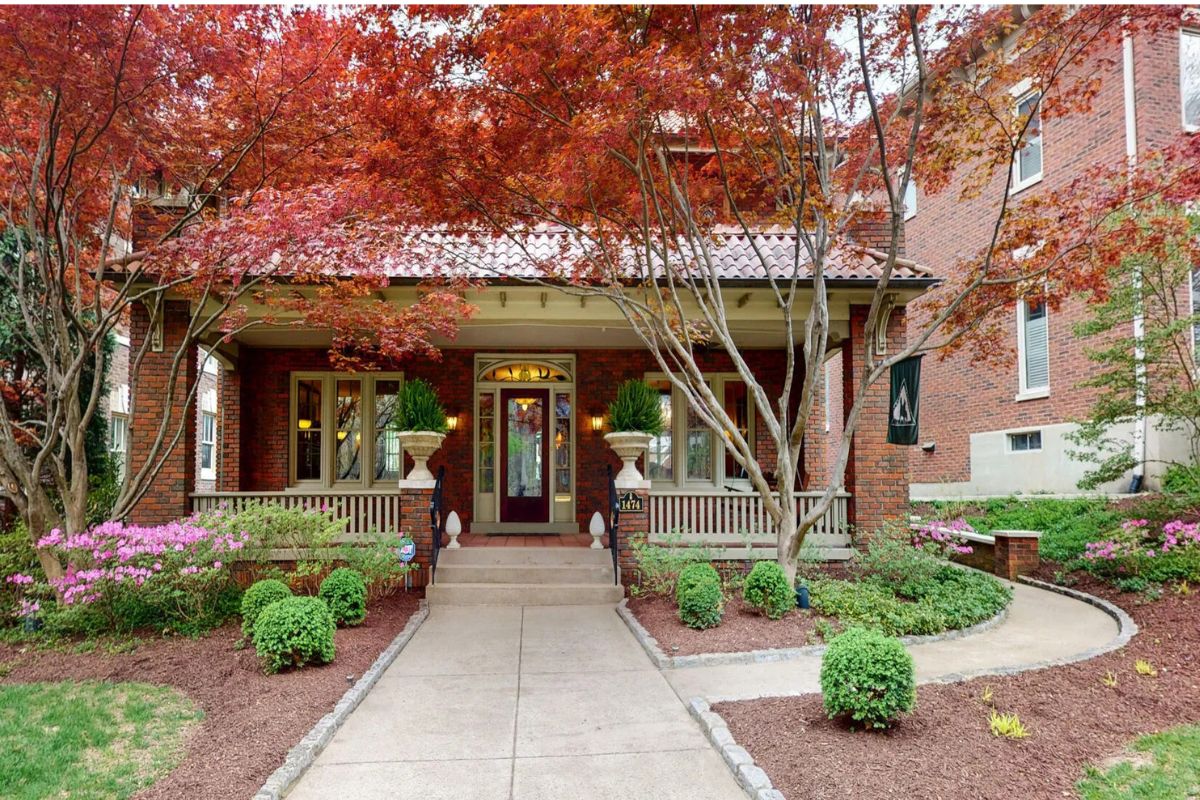 A brick house with a covered front porch on Cherokee Road, Louisville KY, surrounded by landscaped bushes, flowering plants, and red-leafed trees.
