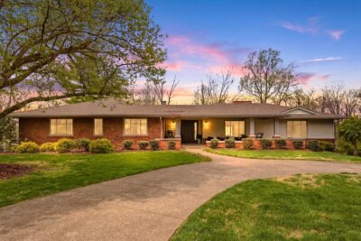 Charming single-story brick house on Mount Rainier Dr in Hills And Dales KY, featuring a circular driveway, lush front lawn, and an evening sky painted with pink and blue hues. 3419.