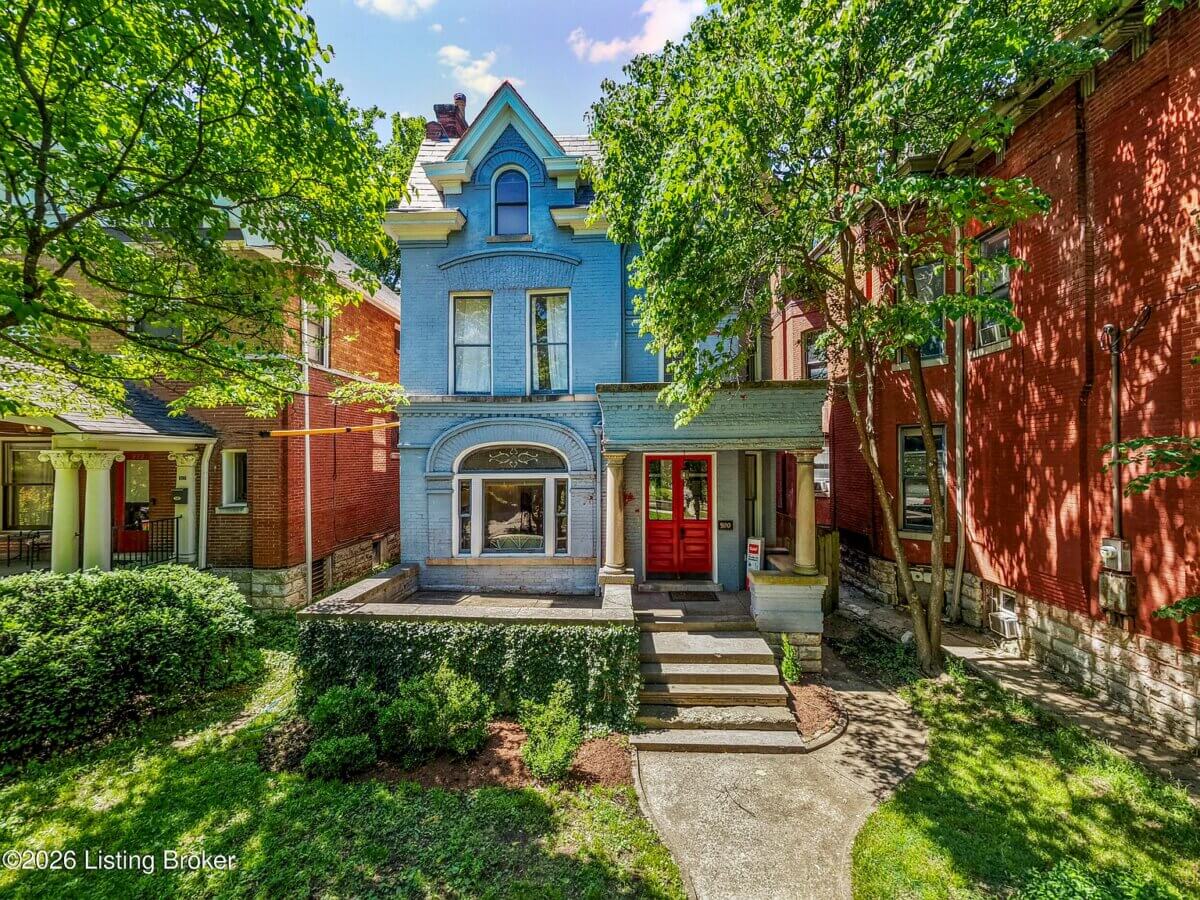 A two-story blue Victorian house with a porch, red front doors, and arched windows sits among mature trees at 920 Cherokee Rd in Louisville KY.