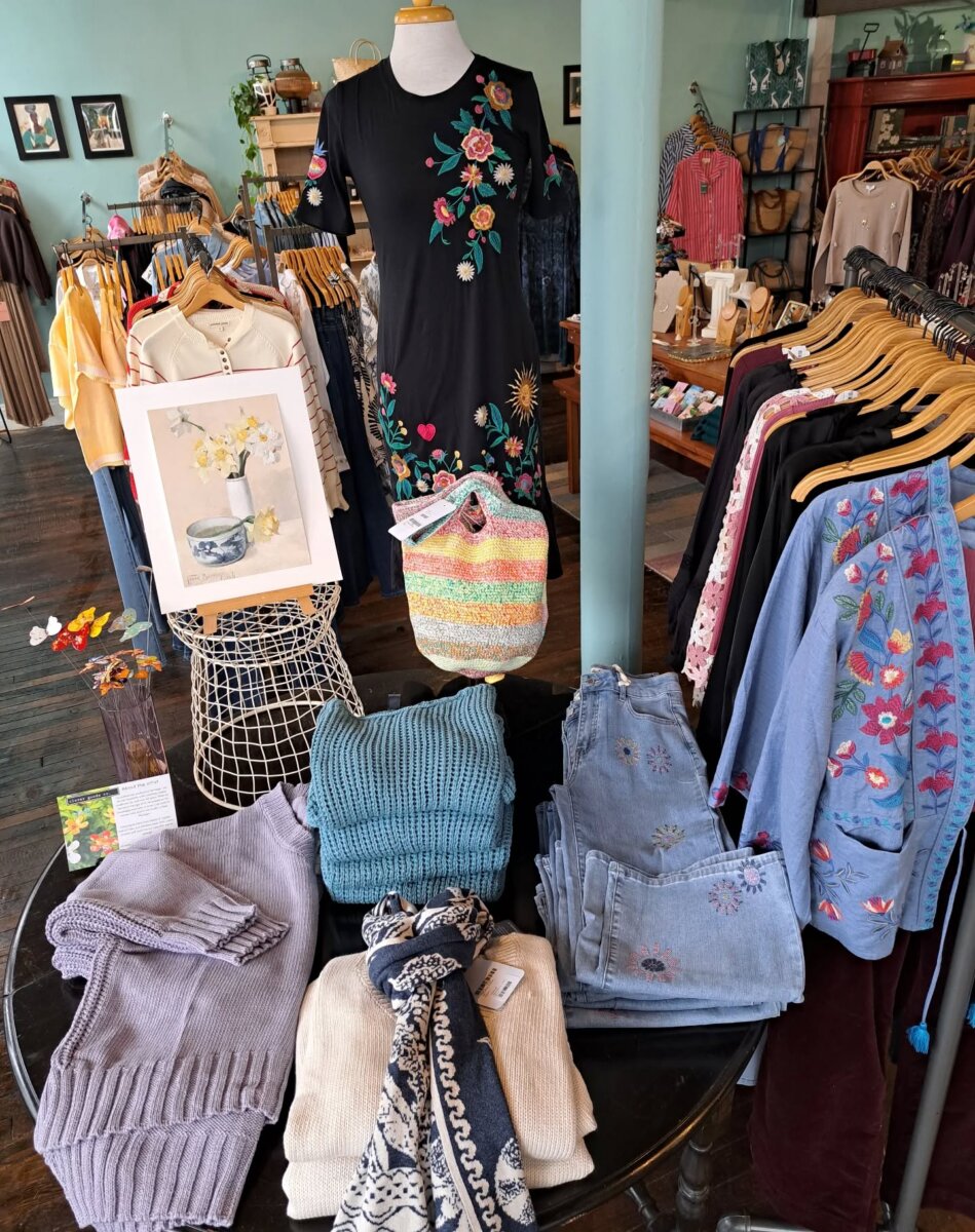 14 Best Women's Boutiques in Louisville - 2 Chic Women's Boutiques in Louisville: A 2026 display with folded sweaters, jeans, scarves, a floral dress on a mannequin, a crocheted bag, and a framed flower illustration arranged on a round table.