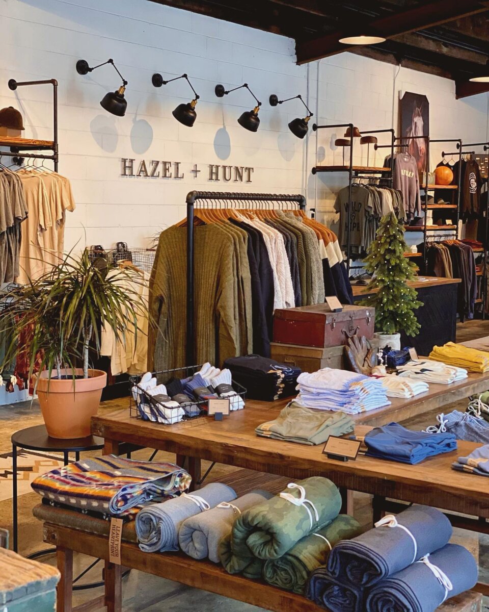 14 Best Women's Boutiques in Louisville - 4 Chic clothing boutique interior with shirts, jackets, and sweaters on racks, folded clothes on tables, a potted plant, a mini Christmas tree, and a "Hazel + Hunt" sign—one of the best boutiques Louisville has to offer.