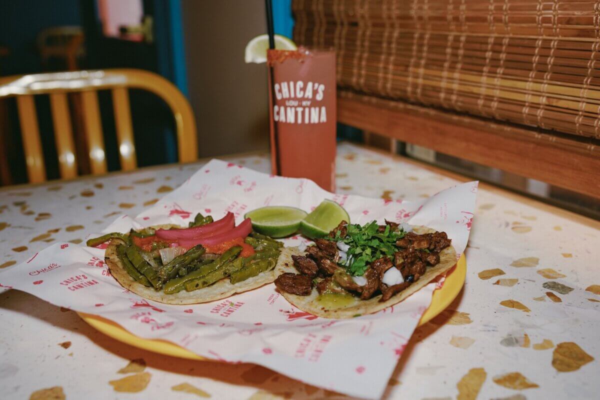 Your Updated Guide to Butchertown - 2 Two tacos on a plate in Butchertown: one with grilled cactus and pickled onions, the other with beef, onions, and cilantro, served with lime wedges—all part of our Updated Guide—with a pink cocktail in the background.