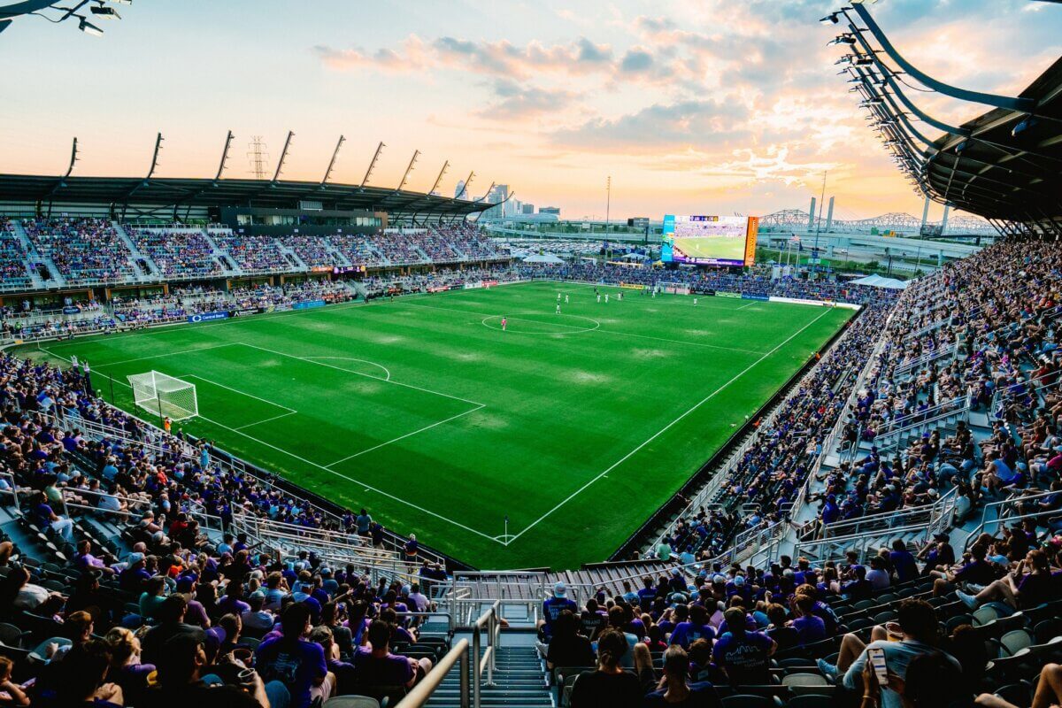 Your Updated Guide to Butchertown - 6 A large soccer stadium in Butchertown filled with spectators during a match at sunset, with players on the field and city buildings in the background—a perfect guide to an unforgettable game day atmosphere.
