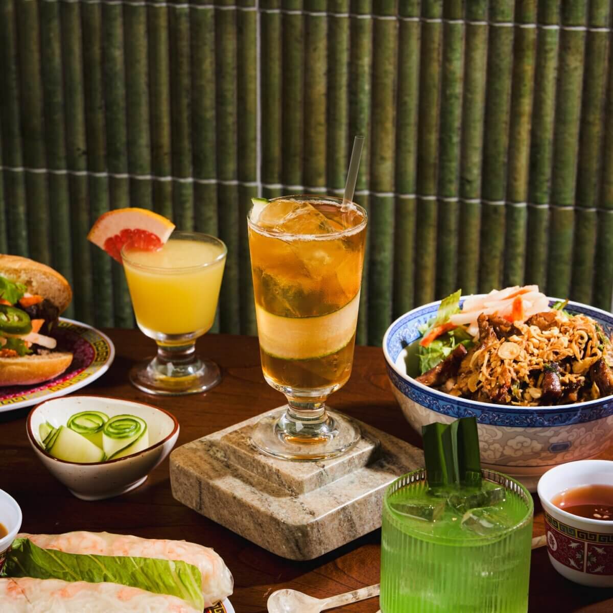 Your Updated Guide to Butchertown - 4 A variety of Vietnamese dishes and drinks on a wooden table in Butchertown, including spring rolls, a banh mi sandwich, rice bowl, sliced cucumbers, and assorted beverages—a delicious scene from our updated guide against a bamboo backdrop.