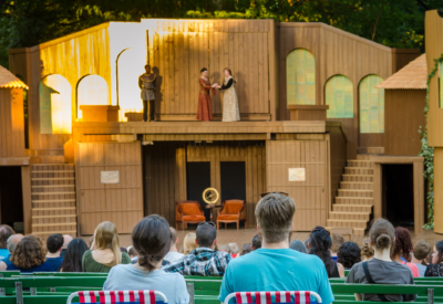 An audience watches an outdoor stage play with four actors performing on a wooden set, featuring two levels and simple props, during daylight.