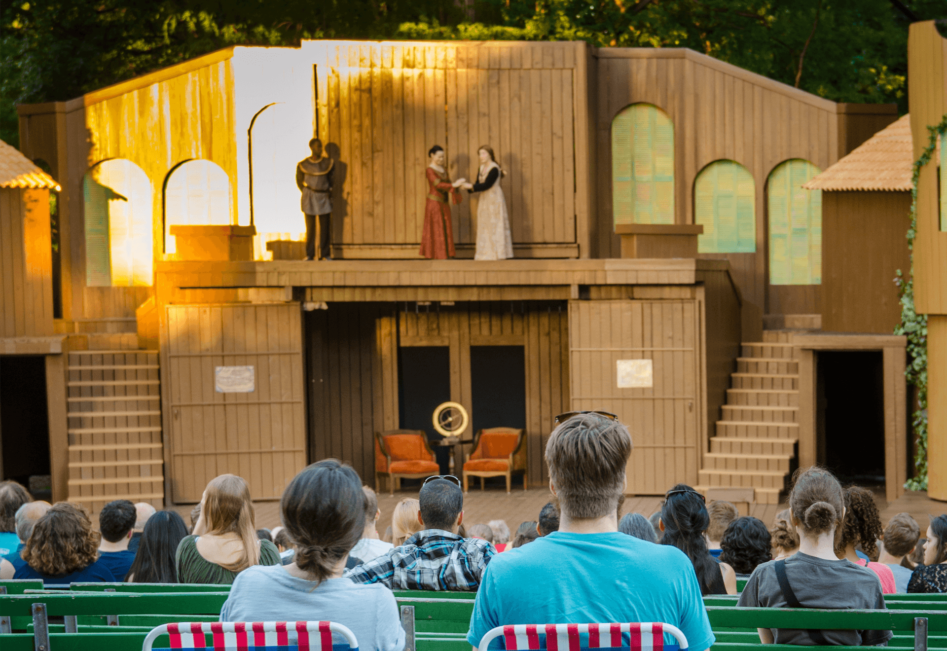 An audience watches an outdoor stage play with four actors performing on a wooden set, featuring two levels and simple props, during daylight.