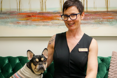 A woman with short dark hair and glasses sits on a green couch next to a French bulldog in a striped sweater, embodying self-care. A colorful painting hangs on the wall behind them.