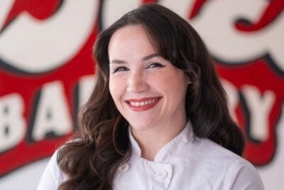 A woman with long brown hair wearing a white chef's coat stands smiling in front of a red and white sign.