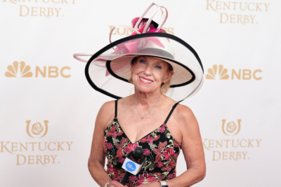 Kirby Adams, Lifestyle Reporter, wearing a large decorative hat and floral dress, holds a microphone on the red carpet at the Kentucky Derby, with NBC and Derby logos in the background—capturing the story of race day style.