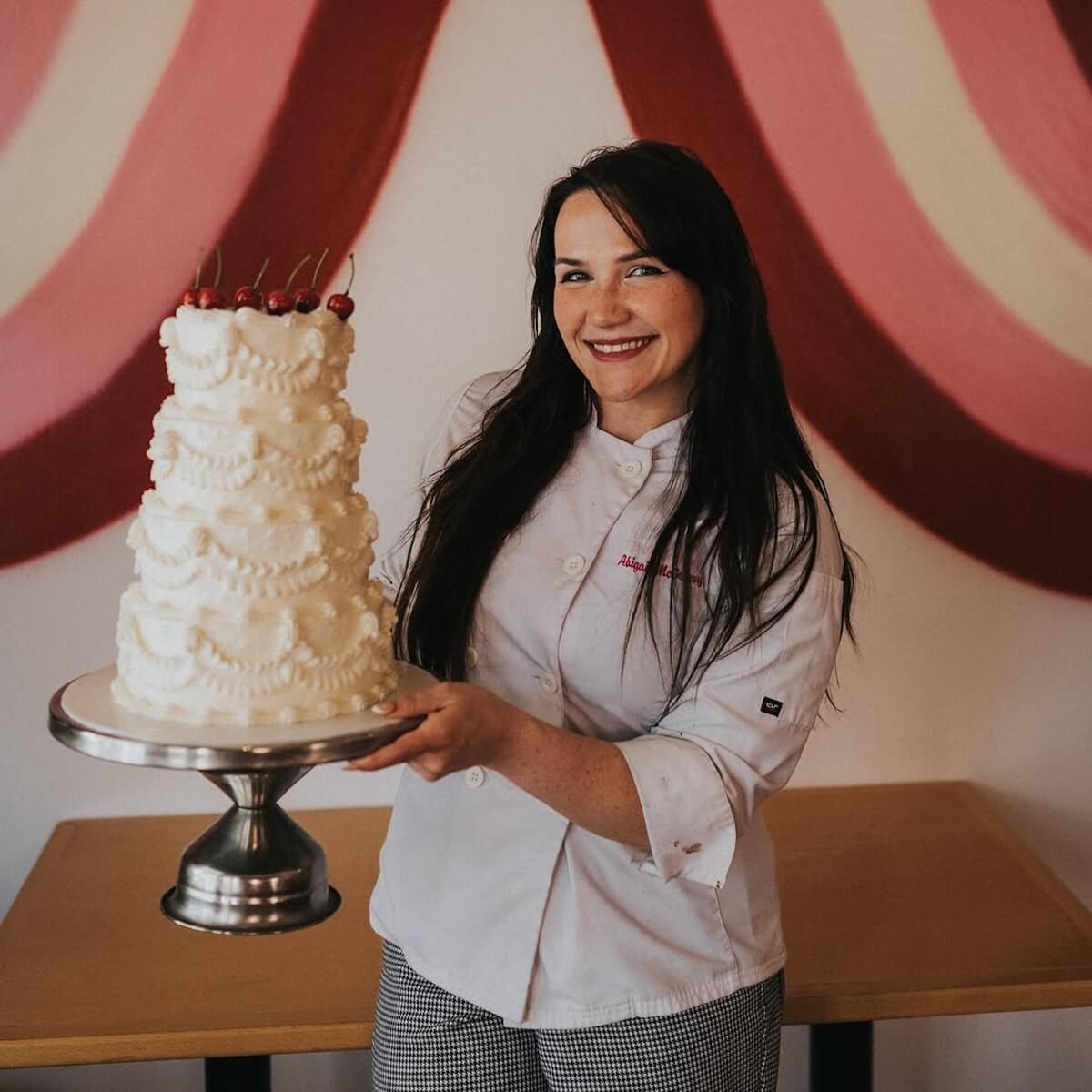 She Didn't Get into Law School. So, She Built a Cookie Empire Instead. - 2 Abigail McGreevy, the woman behind Bae's Baekery, stands in a white chefβs coat holding a tall, ornate white cake topped with cherries, set against a vibrant pink and red wall mural.