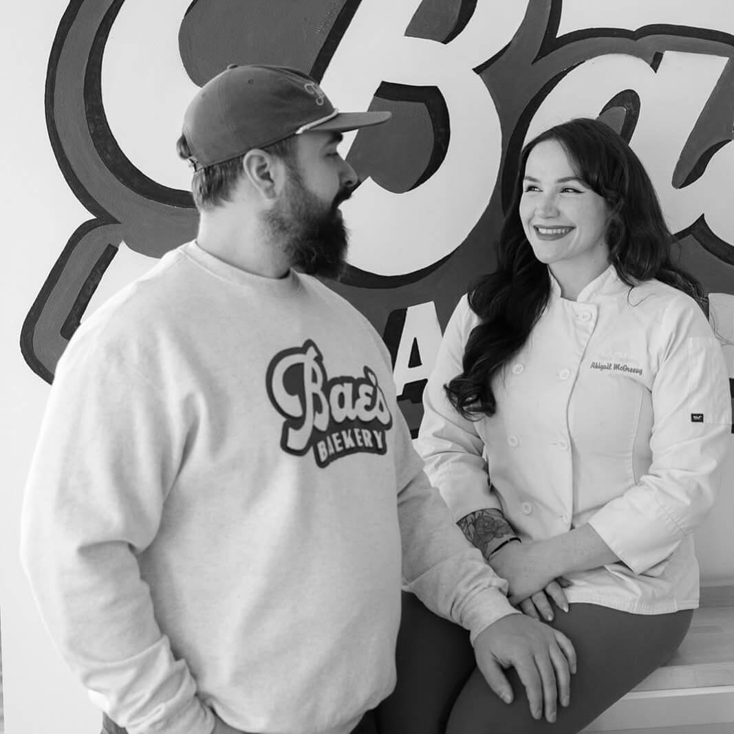 She Didn't Get into Law School. So, She Built a Cookie Empire Instead. - 7 A man in a sweatshirt and Abigail McGreevy, the Woman Behind Bae's Baekery, smile at each other while standing in front of a large "Bae's" sign.