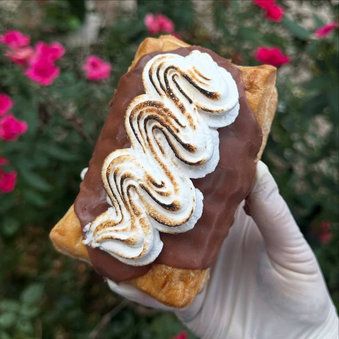 She Didn't Get into Law School. So, She Built a Cookie Empire Instead. - 4 A gloved hand holds a pastry topped with chocolate glaze and swirled toasted meringue from Bae's Baekery, with pink flowers and green foliage in the blurred background.