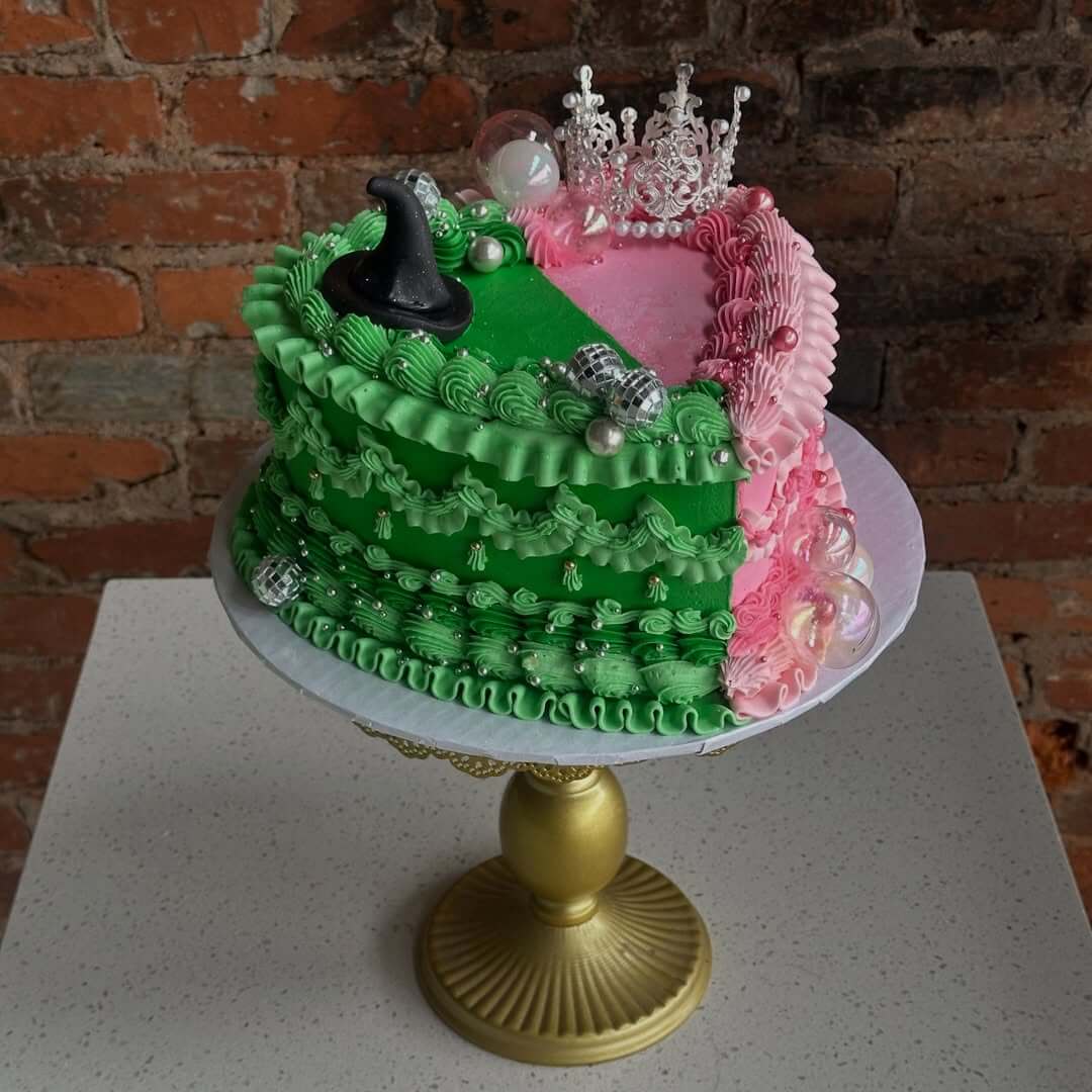 She Didn't Get into Law School. So, She Built a Cookie Empire Instead. - 3 A cake with green and pink frosting split down the middle, designed by Abigail McGreevy of Bae's Baekery, is topped with a black witch hat, silver crown, pearls, and clear bubbles on a gold cake stand.
