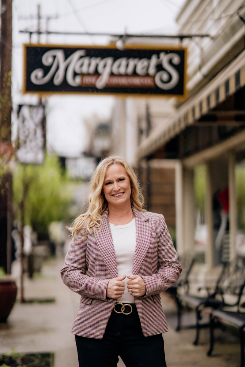 Meet the Woman Behind Margaret’s Fine Consignments - 2 A woman with long blonde hair stands in front of a shop called “Margaret’s,” wearing a plaid blazer, white top, and black pants on a city sidewalk.