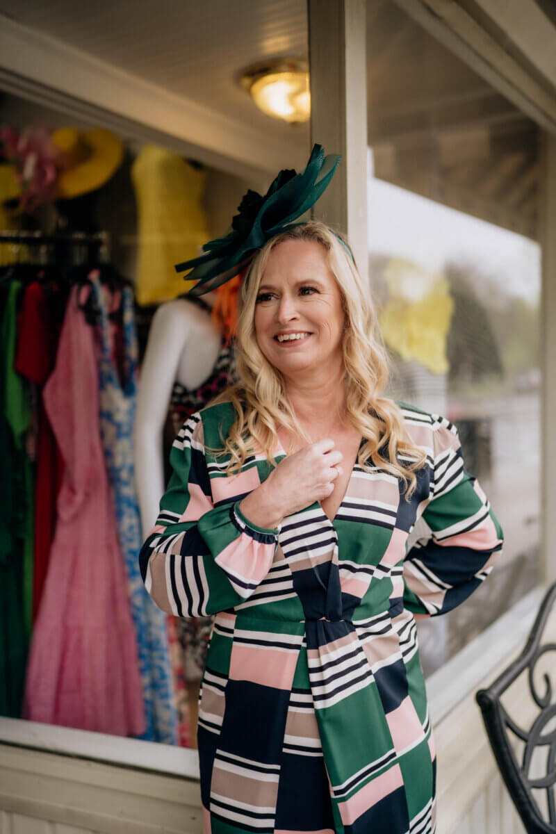 Meet the Woman Behind Margaret’s Fine Consignments - 6 Cara Shelton, with long blonde hair, stands smiling outside a consignment clothing store in a patterned dress and green fascinator, surrounded by the legacy of stylish dresses displayed inside.
