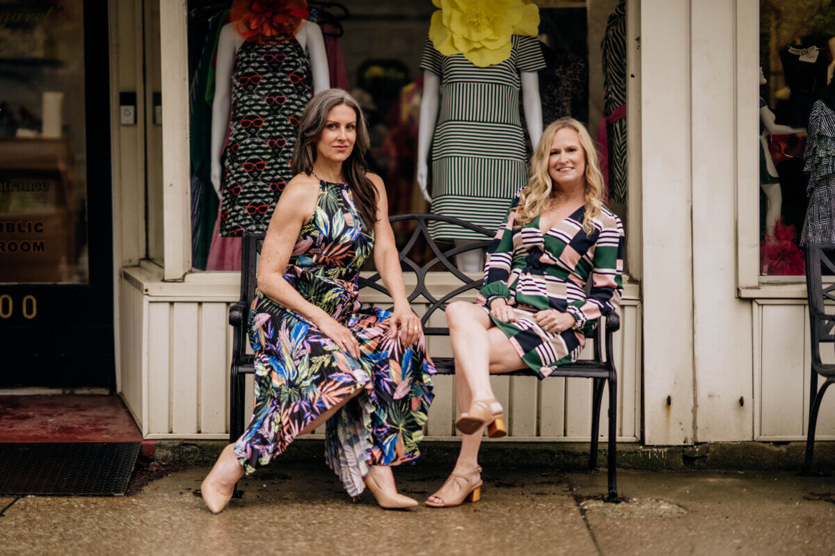 Meet the Woman Behind Margaret’s Fine Consignments - 3 Two women sit on a bench outside a consignment clothing store, both wearing patterned dresses and sandals, with mannequins in similar styles displayed behind them.
