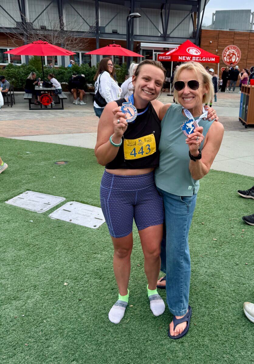 Meet Kirby Adams, the Lifestyle Reporter with a Story of Her Own - 5 Lifestyle Reporter Kirby Adams stands next to another woman, smiling and holding medals—one in athletic gear with race bib 443, the other casually dressed. People and red umbrellas appear in the background.