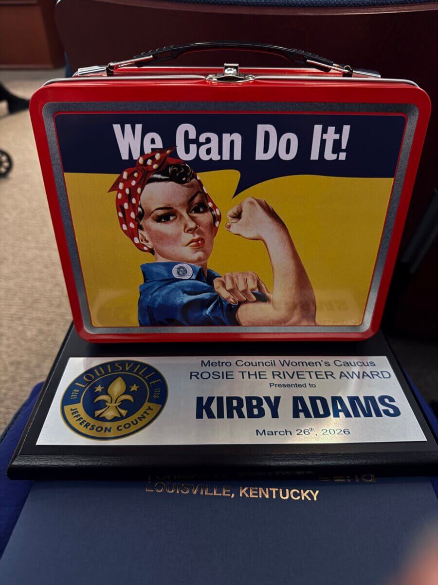 Meet Kirby Adams, the Lifestyle Reporter with a Story of Her Own - 3 A "We Can Do It!" Rosie the Riveter lunchbox sits atop a plaque awarded to Kirby Adams, Lifestyle Reporter, by the Metro Council Women's Caucus in Louisville, Kentucky, on March 26, 2026—a story of empowerment and recognition.