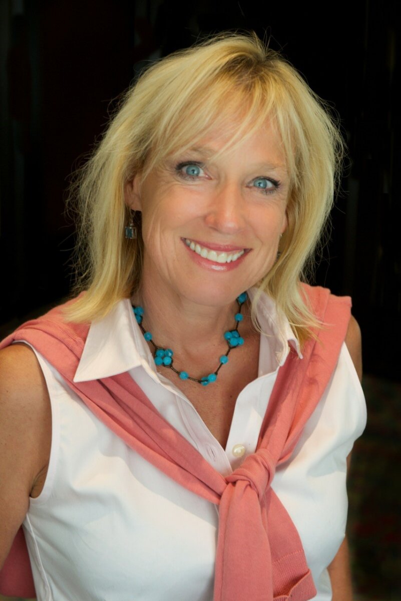Meet Kirby Adams, the Lifestyle Reporter with a Story of Her Own - 2 Smiling woman with blonde hair, Lifestyle Reporter Kirby Adams, wears a white blouse, pink sweater draped over her shoulders, and a turquoise necklace as she stands against a dark background.