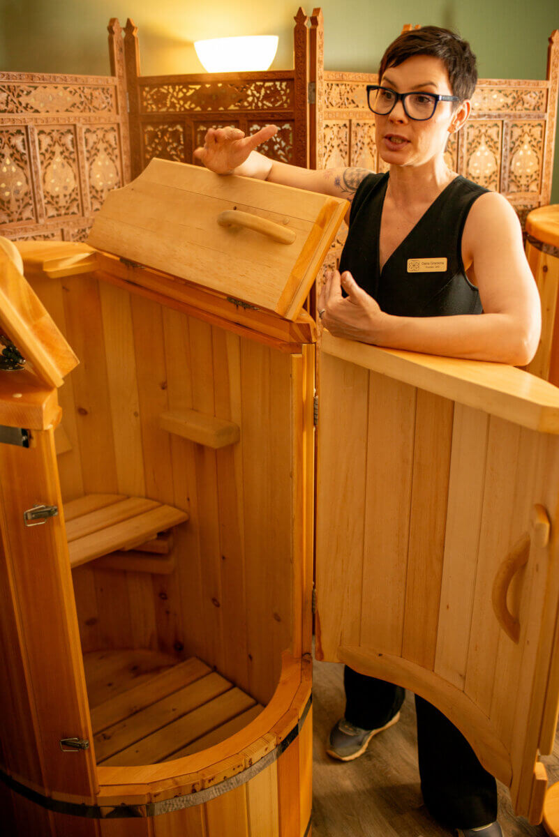 She's Bringing Ancient Wellness Traditions to Louisville - 2 A woman gestures with one hand while standing next to an open wooden sauna pod in a spa-like setting.