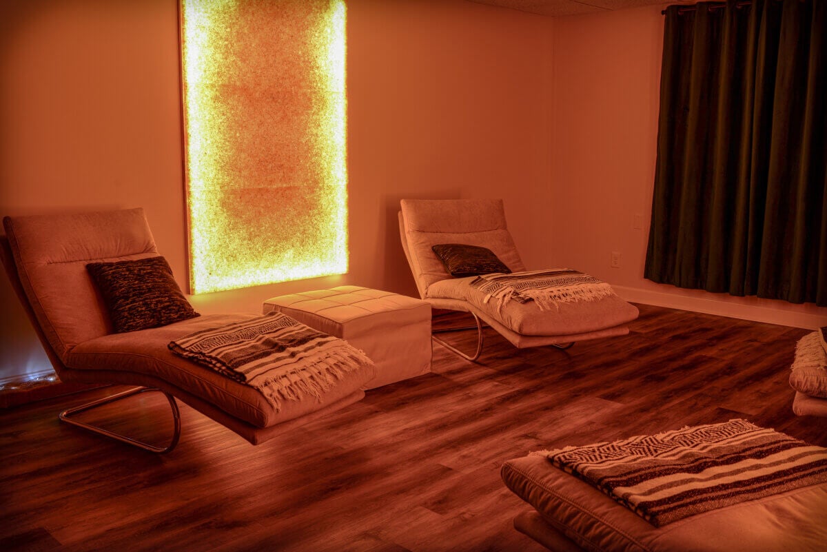 She's Bringing Ancient Wellness Traditions to Louisville - 4 A softly lit room with two modern lounge chairs, a small ottoman, and a glowing wall panel, featuring wood flooring and dark curtains.