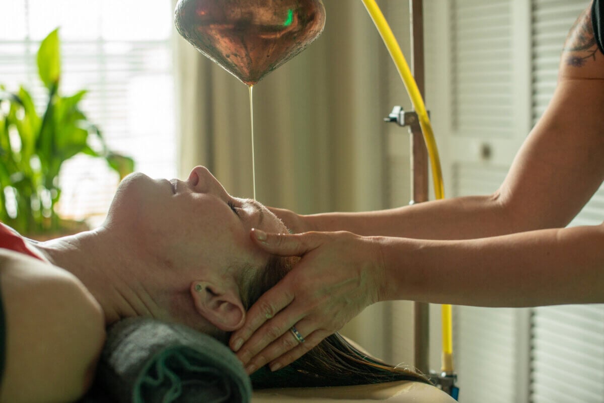 She's Bringing Ancient Wellness Traditions to Louisville - 3 A person receives an Ayurvedic Shirodhara treatment, with oil poured onto their forehead while lying down, as another person supports their head.