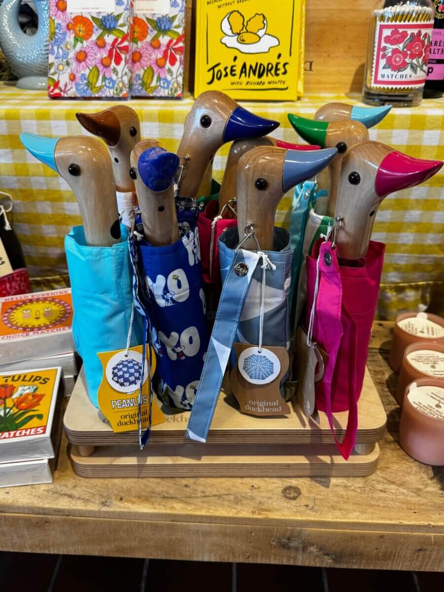 15 Louisville FINDS for May: Spring Fashion, Entertaining Essentials & More - 15 Several umbrella handles shaped like ducks with colorful beaks and matching covers are displayed upright in a wooden stand on a checkered tableclothβa charming Louisville find this May.