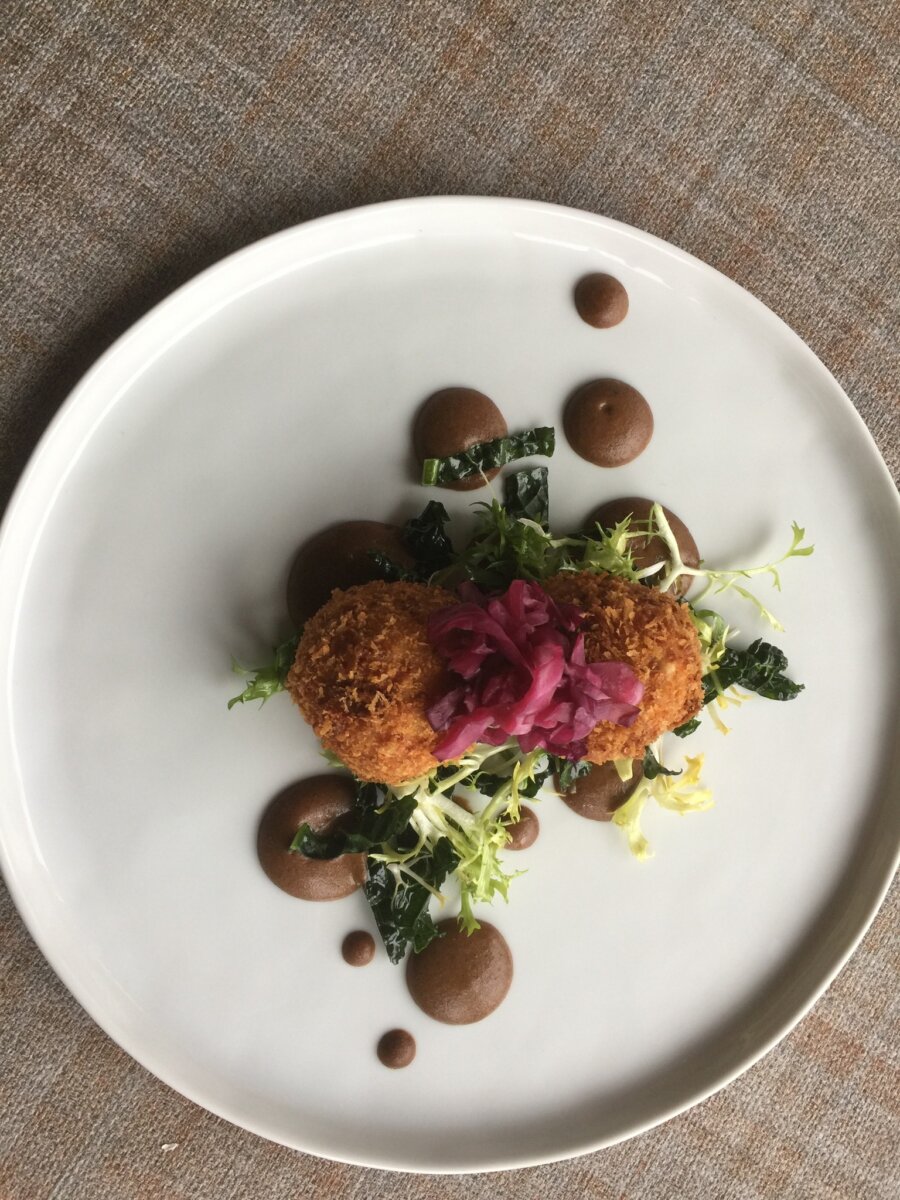 3 New Louisville Restaurants to Try this Month (+2 New Menus!) - 3 Two breaded croquettes are plated with greens, pickled red onions, and dollops of brown sauce on a white plate atop a textured fabric surface.