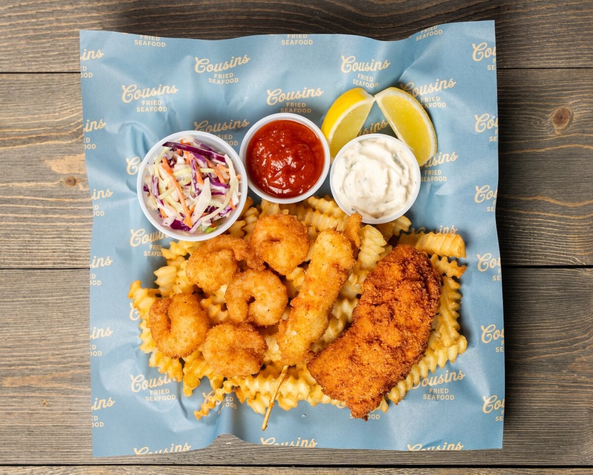 3 New Louisville Restaurants to Try this Month (+2 New Menus!) - 4 A tray with fried shrimp, a breaded fish fillet, crinkle-cut fries, coleslaw, cocktail sauce, tartar sauce, and lemon wedges on branded paper over a wooden surface.